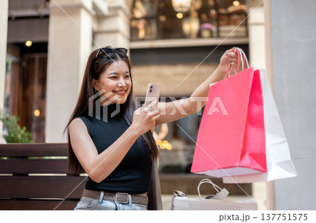 Happy asian woman holding shopping bags taking pics on phone sitting on wooden bench in mall market. Happy asian woman holding shopping bags taking pics on phone sitting on wooden bench in mall market. 137751575