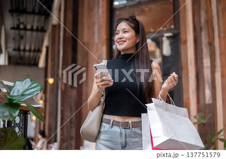 Happy asian woman holding phone carrying shopping bags while walking in front of a cafe in the mall. Happy asian woman holding phone carrying shopping bags while walking in front of a cafe in the mall. 137751579