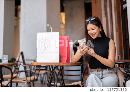 Pretty asian woman holding looking at phone while sitting with shopping bags on wooden table in cafe Pretty asian woman holding looking at phone while sitting with shopping bags on wooden table in cafe 137751586