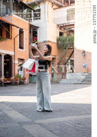 Pretty asian woman carrying shopping bags holding phone taking picture while standing in mall plaza. Pretty asian woman carrying shopping bags holding phone taking picture while standing in mall plaza. 137751595