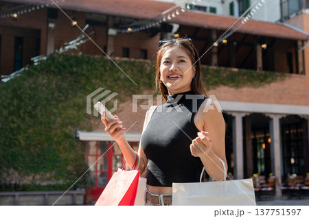Pretty asian woman carrying shopping bag holding phone while standing or walking in brick wall plaza Pretty asian woman carrying shopping bag holding phone while standing or walking in brick wall plaza 137751597