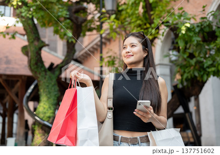 Pretty asian woman holding smartphone carrying shopping bags while standing or walking in mall plaza Pretty asian woman holding smartphone carrying shopping bags while standing or walking in mall plaza 137751602