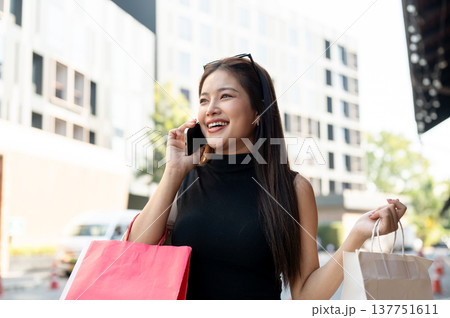 Pretty asian woman talking on phone and carrying shopping bags while standing or walking by street. 137751611