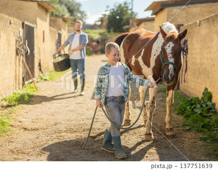 Happy boy leads a horse by the bridle for a walk in pasture Happy boy leads a horse by the bridle for a walk in pasture 137751639
