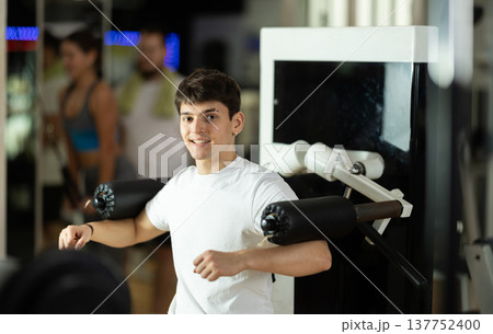 Guy in gym performs exercises for arm muscles using simulators and fitness center equipment. Guy in gym performs exercises for arm muscles using simulators and fitness center equipment. 137752400