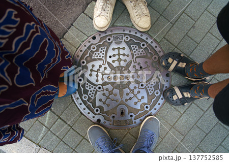 Tokyo, Japan - Oct 14 2024, close-up view of a metal manhole cover with a lotus flower depicted around it, with legs of a family of four standing around it, Ginza district, Tokyo, Japan 137752585