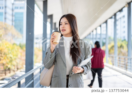 Asian woman student office worker drinking coffee holding tablet while walking on sun lit footbridge 137753044