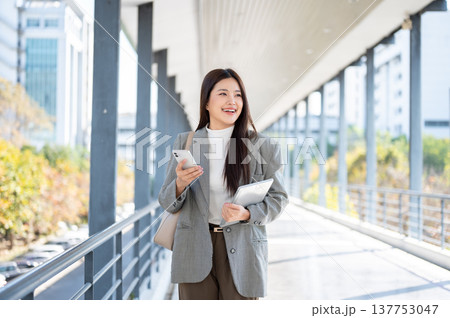 Happy asian woman student office worker holding tablet and phone while walking on sun lit footbridge 137753047
