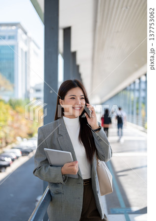 Asian woman student office worker talking on phone holding tablet as standing walking on footbridge. Asian woman student office worker talking on phone holding tablet as standing walking on footbridge. 137753062