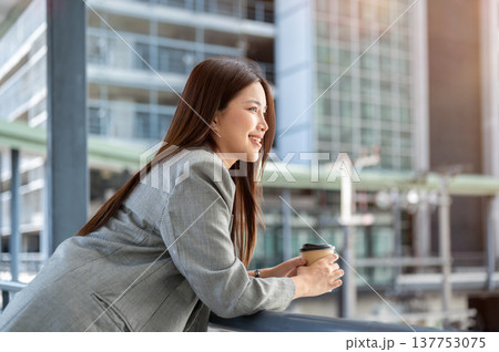 Asian woman office worker in suit holding coffee cup leaning over a railing on bridge with cityscape 137753075