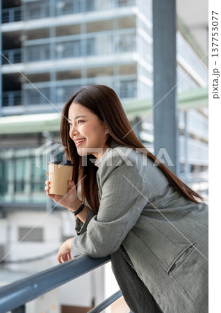 Asian woman office worker holding cup drinking coffee leaning over railing of bridge with cityscape. 137753077