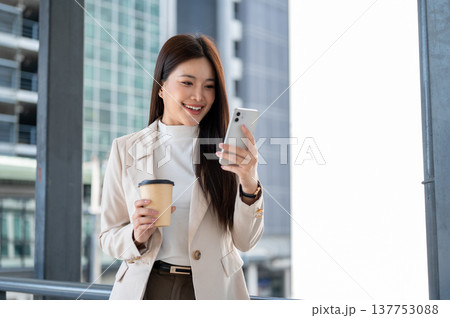 Happy asian woman office worker holding coffee looking at phone as standing on bridge with cityscape 137753088