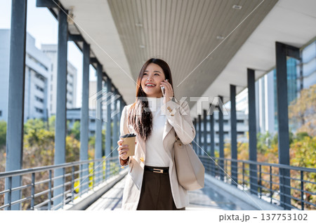 Pretty asian woman office worker holding coffee talking on phone as walking standing on a footbridge Pretty asian woman office worker holding coffee talking on phone as walking standing on a footbridge 137753102