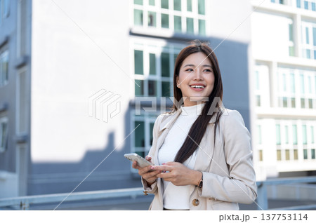 Pretty asian woman in beige coat holding phone looking away while standing or walking aside building 137753114