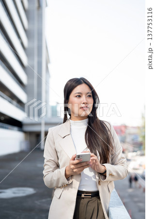 Asian woman in beige blazer holding phone looking away while standing walking aside building railing 137753116