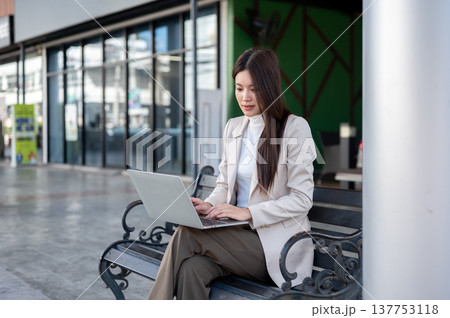 Pretty asian woman in beige coat using laptop while sitting crossed legs on metal bench at sidewalk. Pretty asian woman in beige coat using laptop while sitting crossed legs on metal bench at sidewalk. 137753118