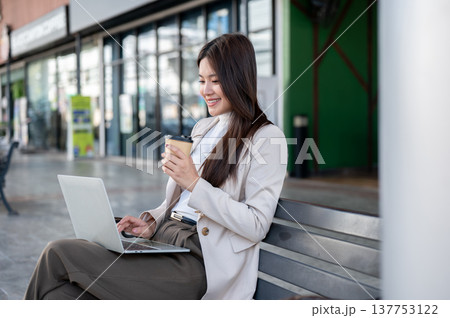 Pretty asian woman in beige coat holding coffee looking at laptop while sitting on bench at sidewalk 137753122
