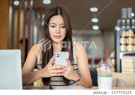 Pretty asian woman holding looking at phone aside laptop and coffee while sitting at table in cafe. Pretty asian woman holding looking at phone aside laptop and coffee while sitting at table in cafe. 137753125