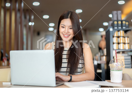 Smiling asian woman looking and typing on laptop while sitting with book and coffee at table in cafe Smiling asian woman looking and typing on laptop while sitting with book and coffee at table in cafe 137753134