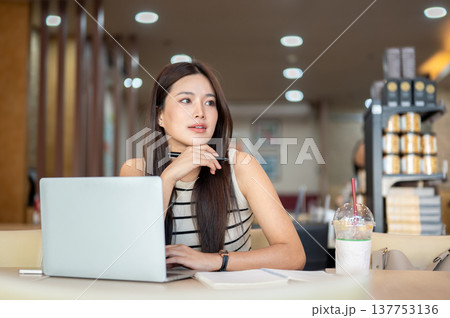 Asian woman holding pen under chin thinking over laptop with book and coffee sits at table in cafe. Asian woman holding pen under chin thinking over laptop with book and coffee sits at table in cafe. 137753136