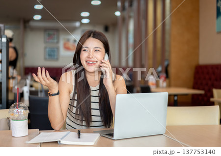 Smiling asian woman talking on phone with one hand raised over laptop and book sits at table in cafe Smiling asian woman talking on phone with one hand raised over laptop and book sits at table in cafe 137753140
