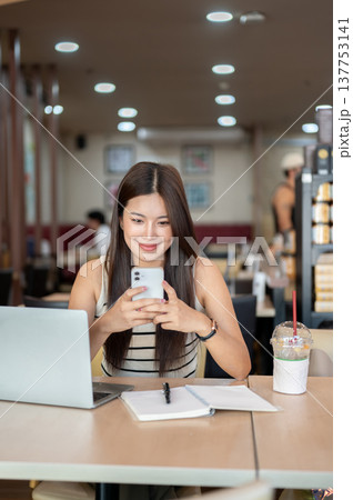 Asian woman holding phone with both hands over laptop aside book and coffee sitting at table in cafe Asian woman holding phone with both hands over laptop aside book and coffee sitting at table in cafe 137753141