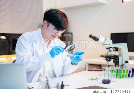 Scientist researcher man holds magnifying glass looking at test sample dish aside microscope in lab. 137753418