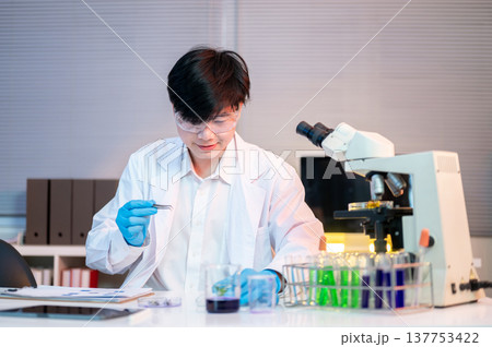 Scientist researcher man wears gloves holding forceps looking at test sample on working table in lab Scientist researcher man wears gloves holding forceps looking at test sample on working table in lab 137753422