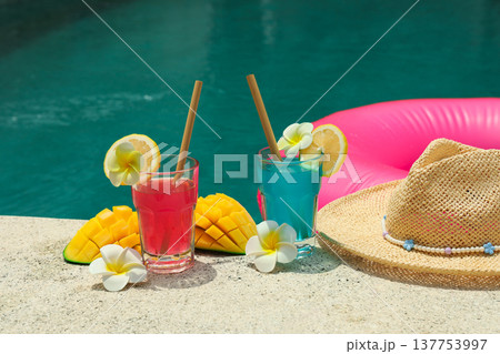 Two colorful drinks sit by a pool with fruit and flowers 137753997