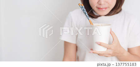 Happy young Asian woman enjoying a delicious cup of hot instant noodles while relaxing at home, Cheerful female eating instant ramen with a smile at apartment. Simple lifestyle and quick meal concept Happy young Asian woman enjoying a delicious cup of hot instant noodles while relaxing at home, Cheerful female eating instant ramen with a smile at apartment. Simple lifestyle and quick meal concept 137755183
