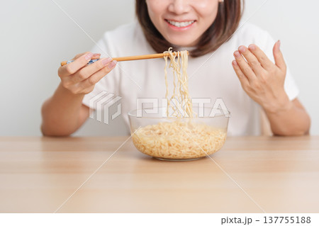 Happy young Asian woman enjoying a delicious bowl of hot instant noodles while relaxing at home, Cheerful female eating instant ramen with a smile at apartment. Simple lifestyle and quick meal concept 137755188