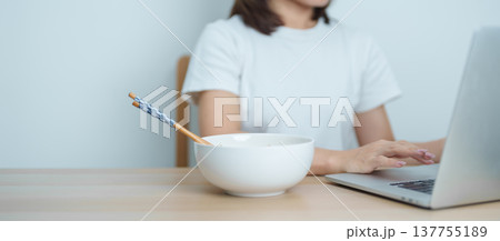Happy young Asian woman working late at night on laptop while enjoying a bowl of hot instant ramen noodles at home. Cheerful female having a quick meal during work from home. Simple lifestyle concept Happy young Asian woman working late at night on laptop while enjoying a bowl of hot instant ramen noodles at home. Cheerful female having a quick meal during work from home. Simple lifestyle concept 137755189