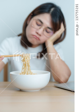 Bored young Asian woman working late at night on laptop while a bowl of hot instant ramen noodles at home. Unhappy female having a quick meal during work from home. Simple lifestyle concept 137755191