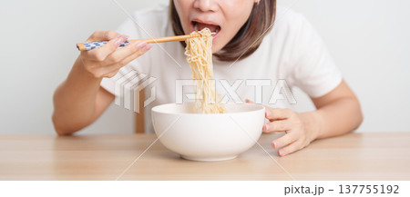 Happy young Asian woman enjoying a delicious bowl of hot instant noodles while relaxing at home, Cheerful female eating instant ramen with a smile at apartment. Simple lifestyle and quick meal concept 137755192