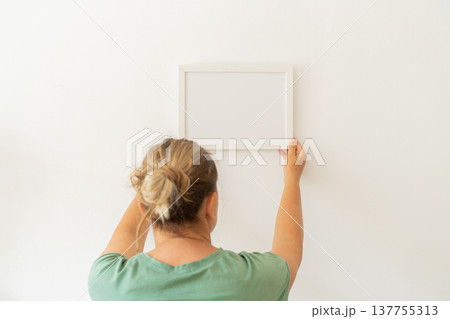 Blonde woman in green t-shirt hanging empty picture frame on white wall at home. Modern minimalist mockup, empty template for text Blonde woman in green t-shirt hanging empty picture frame on white wall at home. Modern minimalist mockup, empty template for text 137755313