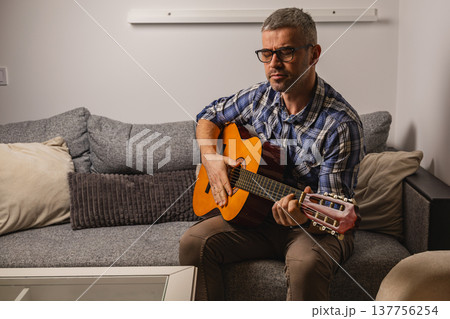 Man plays guitar while sitting on sofa during evening indoors Man plays guitar while sitting on sofa during evening indoors 137756254