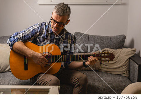 Man plays guitar while sitting on couch in modern living room 137756258