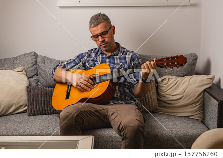 Man plays guitar while sitting on couch in living room Man plays guitar while sitting on couch in living room 137756260