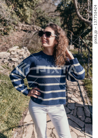 Woman striped sweater standing on stone path in sunny garden wearing sunglasses during bright afternoon Woman striped sweater standing on stone path in sunny garden wearing sunglasses during bright afternoon 137758424