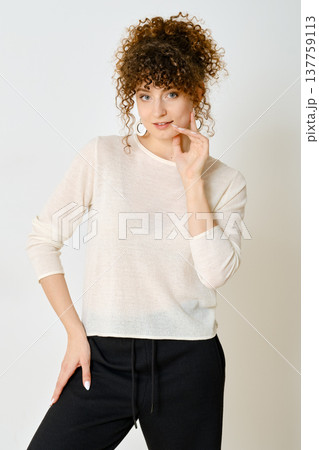 Curly-haired model posing in a light pullover and black pants against white wall 137759113