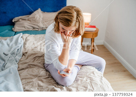 Concerned woman reading text on smartphone while sitting on bed. Concerned woman reading text on smartphone while sitting on bed. 137759324
