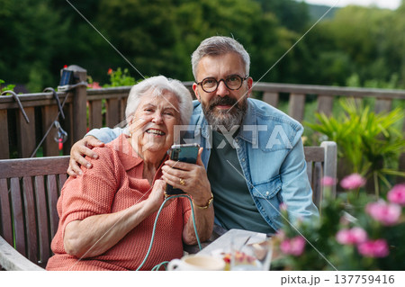 Mature son spending time with elderly mother, showing her something on smartphone. Mature son spending time with elderly mother, showing her something on smartphone. 137759416
