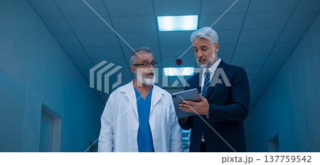Manager walking with doctor down the hallway, showing him document in tablet. Manager walking with doctor down the hallway, showing him document in tablet. 137759542