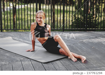 Preteen girl sitting on yoga mat in sportswear outdoors in forest after fitness training, smiling and looking at camera. Healthy child lifestyle, active recreation in nature, Athletic kid Preteen girl sitting on yoga mat in sportswear outdoors in forest after fitness training, smiling and looking at camera. Healthy child lifestyle, active recreation in nature, Athletic kid 137761294