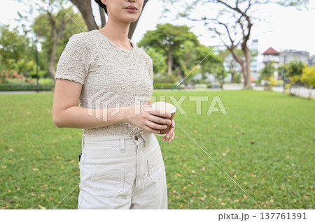 Calm young woman standing in green park holding cup of coffee. relaxing outdoor lifestyle moment enjoying peaceful break during daytime 137761391
