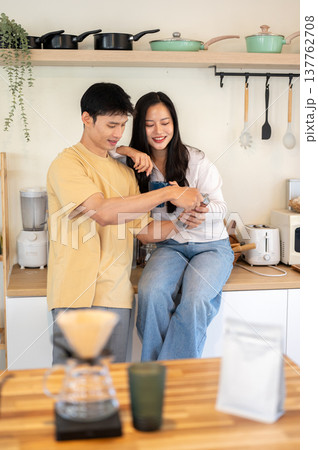 Asian couple woman sits on counter and man looking at powder in grinder as making coffee in kitchen. Asian couple woman sits on counter and man looking at powder in grinder as making coffee in kitchen. 137762708