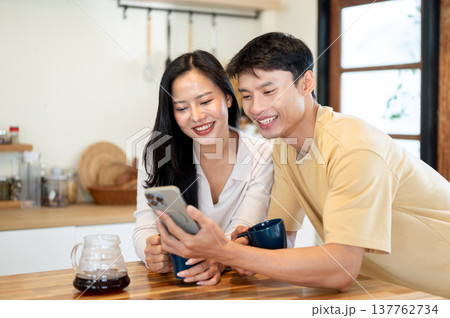 Pretty asian woman and man couple looking at phone watching videos making coffee at kitchen counter. Pretty asian woman and man couple looking at phone watching videos making coffee at kitchen counter. 137762734