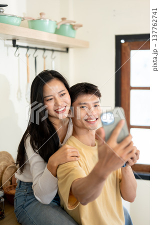 Pretty asian woman and man couple holding phone taking picture as sitting leaning on kitchen counter Pretty asian woman and man couple holding phone taking picture as sitting leaning on kitchen counter 137762741
