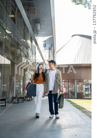 Full body asian man woman couple holding hands and shopping bags while walking on pavement in plaza. Full body asian man woman couple holding hands and shopping bags while walking on pavement in plaza. 137762852