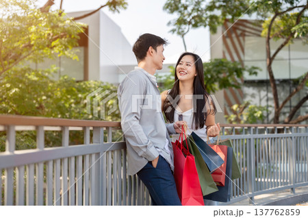 Asian couple a man holding shopping bags talking to a smiling woman while standing on bridge in park 137762859